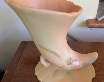 May include: A pale pink ceramic vase in the shape of a horn with a floral design. The vase has a wide opening and a rounded base.