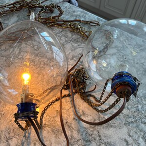 Op de afbeelding: Twee heldere glazen globe hanglampen met zichtbare gloeilampen. De lampen hebben blauwe en bruine accenten, met metalen kettingen en bruine snoeren. De lampen hangen aan het plafond.