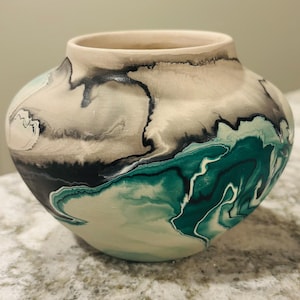 Op de afbeelding: Een ronde keramische vaas met een matte afwerking en een gemarmerd patroon in tinten teal, zwart en crème. De vaas heeft een brede, bolvormige body die taps toeloopt naar een ronde opening aan de bovenkant, met vloeiende, organische kleurwervelingen.