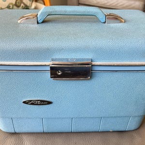 May include: A light blue vintage Forecast travel case with a silver handle and lock. The case has a textured surface and a silver trim around the top and bottom.