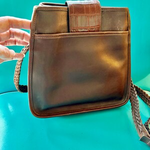 Puede incluir: Un bolso de hombro de cuero marr&oacute;n con una correa trenzada y una solapa con estampado de cocodrilo.