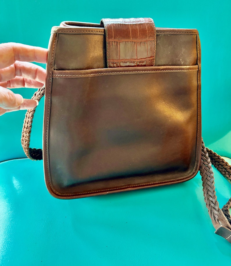 Puede incluir: Un bolso de hombro de cuero marr&oacute;n con una correa trenzada y una solapa con estampado de cocodrilo.