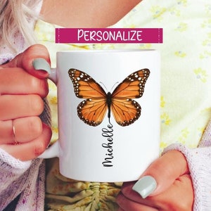 Peut inclure: Une tasse en c&eacute;ramique blanche avec un nom personnalis&eacute;, "Michelle", imprim&eacute; en &eacute;criture noire. Un grand papillon orange avec des marques noires est centr&eacute; sur la tasse.
