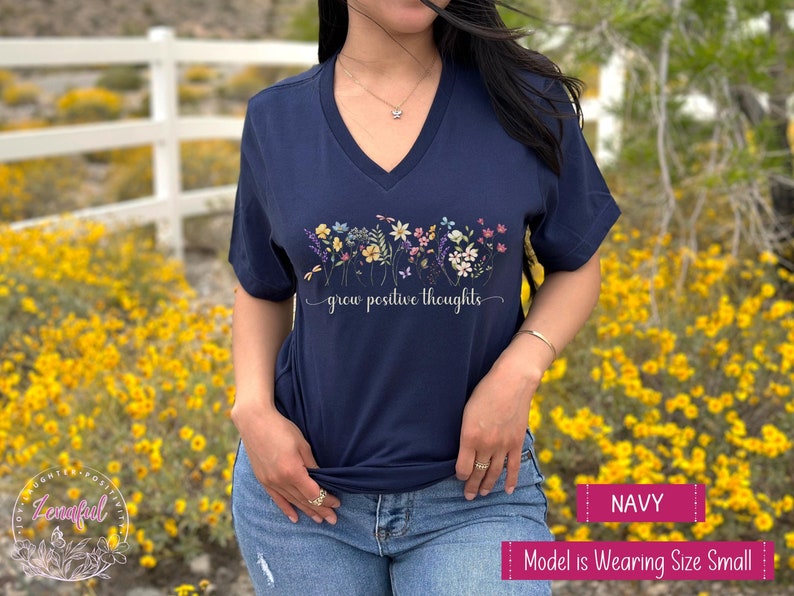 May include: A navy blue v-neck t-shirt with a floral design and the text 'grow positive thoughts' printed on the front. The shirt is displayed on a model wearing blue jeans.