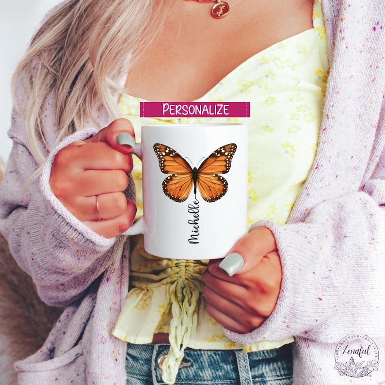 Peut inclure: Une tasse en c&eacute;ramique blanche avec un nom personnalis&eacute;, "Michelle", imprim&eacute; en &eacute;criture noire. Un grand papillon orange avec des marques noires est centr&eacute; sur la tasse.