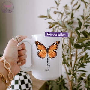 Peut inclure: Une tasse en c&eacute;ramique blanche avec un motif de papillon monarque et le nom "Suzanna" &eacute;crit verticalement en dessous. La tasse est tenue par une main.