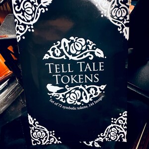 May include: A black box with white decorative corners and the text "TELL TALE TOKENS" in white. The box has the text "Set of 72 symbolic tokens. 144 Images." in white.