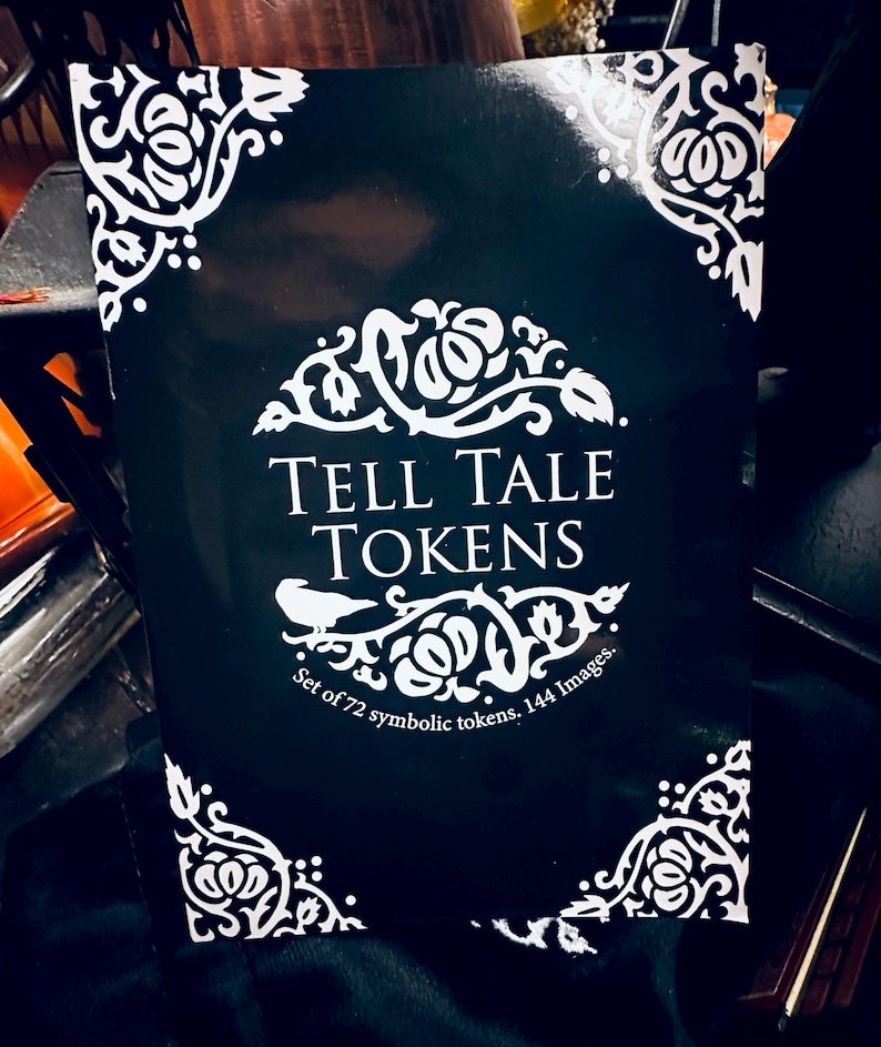 May include: A black box with white decorative corners and the text "TELL TALE TOKENS" in white. The box has the text "Set of 72 symbolic tokens. 144 Images." in white.