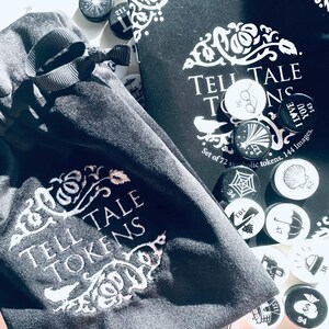 May include: A black velvet bag with a white embroidered design that says "Tell Tale Tokens". The bag is open and there are small round tokens inside. The tokens are black and white and have various designs, including a spider web, a seashell, a pair of glasses, a star, and a shoe.