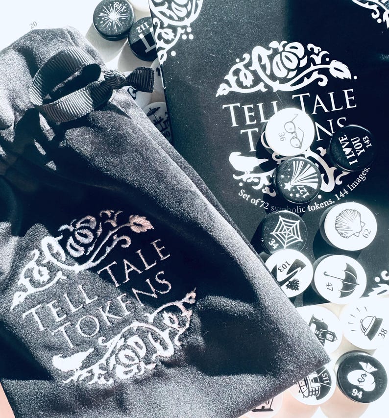 May include: A black velvet bag with a white embroidered design that says "Tell Tale Tokens". The bag is open and there are small round tokens inside. The tokens are black and white and have various designs, including a spider web, a seashell, a pair of glasses, a star, and a shoe.