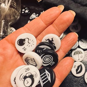 May include: A hand holding a variety of black and white game pieces. The pieces feature different symbols, including a bomb, a spiral, a building, and an alarm clock. The numbers 16, 20, 120, and 121 are visible on the pieces.