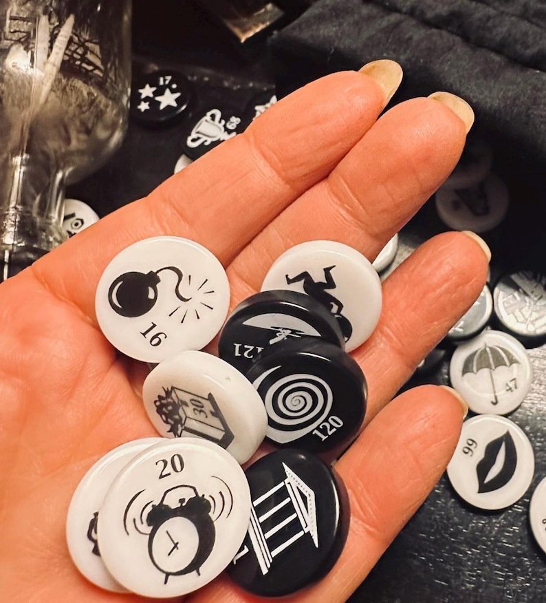 May include: A hand holding a variety of black and white game pieces. The pieces feature different symbols, including a bomb, a spiral, a building, and an alarm clock. The numbers 16, 20, 120, and 121 are visible on the pieces.