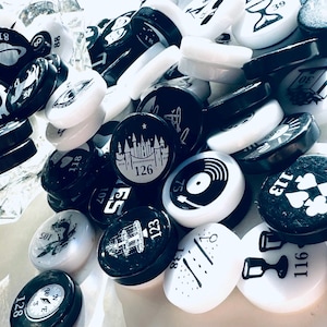 May include: A collection of black and white round tokens with various symbols and numbers. The tokens are made of a smooth, glossy material and have a raised design. Some of the numbers on the tokens include 39, 30, 113, 116, 75, 88, 126, 138, 123, 107, 81, 118, 102, 105, 128.