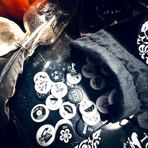 May include: A black velvet bag with a white drawstring sits on a black surface. The bag is filled with round, white tokens with black symbols and numbers. The tokens include symbols like a key, a star, a record player, a flower, a lipstick, and an umbrella. The tokens also have numbers on them, such as 107, 138, and 143.