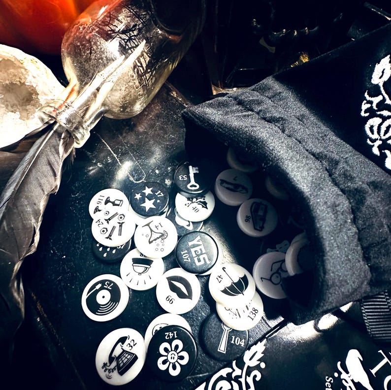 May include: A black velvet bag with a white drawstring sits on a black surface. The bag is filled with round, white tokens with black symbols and numbers. The tokens include symbols like a key, a star, a record player, a flower, a lipstick, and an umbrella. The tokens also have numbers on them, such as 107, 138, and 143.