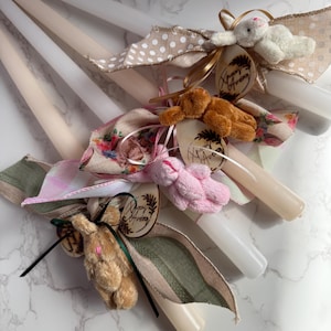 May include: A collection of Easter candles, each adorned with a plush bunny, decorative ribbons, and wooden tags. The candles vary in color, with white, cream, and off-white hues. Ribbons feature polka dots, floral patterns, and solid colors.