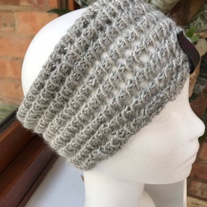 May include: A gray knitted headband with a textured, open weave design. The headband is made of a soft, wool-like material and features a small, dark brown leather tag on the side. The headband is displayed on a white mannequin head.