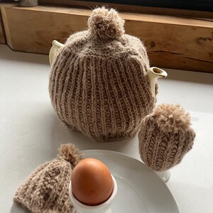 May include: A beige knitted tea cozy with a pom-pom on top, a matching egg cozy, and two egg cup cozies. A brown egg sits in a white egg cup on a white plate with a spoon. The tea cozy covers a white teapot.