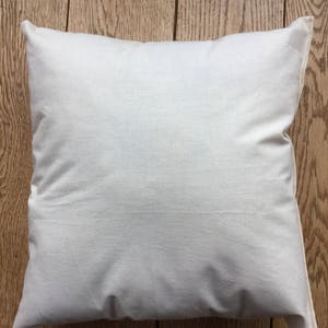 May include: A square throw pillow cover made of plain, off-white, unbleached cotton fabric. The pillow cover features a simple, seamless design with a visible texture characteristic of natural woven cotton fibers, presented in a clean, neutral tone.