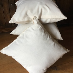 May include: A stack of three square, off-white throw pillows made of a plain, solid-colored fabric. Each pillow features a simple, clean design with visible seams along the edges, presented in a stacked arrangement with a smooth, matte texture.