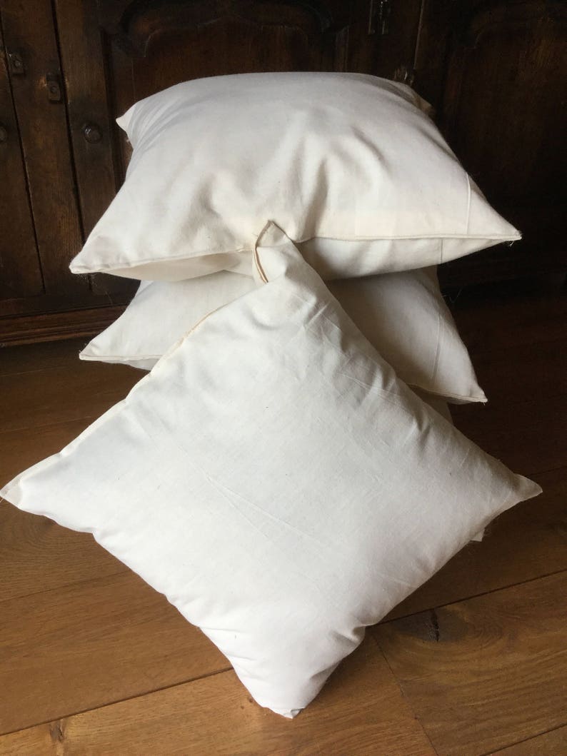 May include: A stack of three square, off-white throw pillows made of a plain, solid-colored fabric. Each pillow features a simple, clean design with visible seams along the edges, presented in a stacked arrangement with a smooth, matte texture.