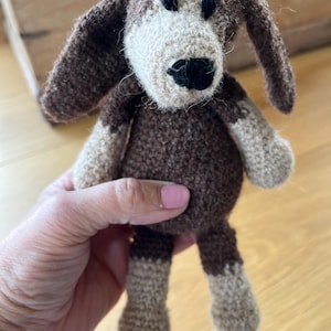 May include: A handmade crochet dog toy in shades of brown and beige. The dog has long floppy ears, a black nose, and is being held by a person. The toy is a cute, soft, and cuddly item.