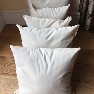 May include: A series of four square, off-white, plain fabric throw pillows arranged in a single line, one behind the other. Each pillow features a simple, unadorned, solid-colored textile cover with visible seams along the edges and a soft, plush appearance.