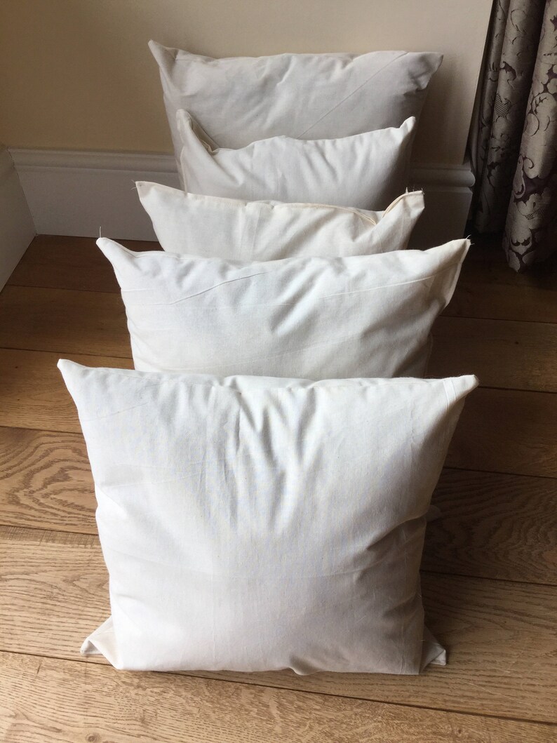 May include: A series of four square, off-white, plain fabric throw pillows arranged in a single line, one behind the other. Each pillow features a simple, unadorned, solid-colored textile cover with visible seams along the edges and a soft, plush appearance.