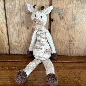 May include: A handmade crochet giraffe plush toy. The giraffe is primarily off-white with brown accents on the face, hooves, and spots. It has floppy ears and long legs, giving it a whimsical appearance. The toy is sitting on a wooden surface.