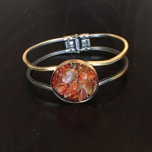Pode incluir: Um bracelete de ouro e prata com uma incrustação redonda de pedra de cornalina. A pedra de cornalina está cheia de pequenas pedras vermelhas, laranja e transparentes.
