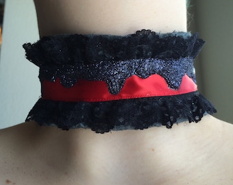May include: Black lace choker with a red satin ribbon and a glittery top layer. The choker has a ruffled edge and is worn around the neck. The design is gothic and elegant.
