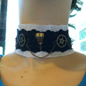 May include: A black and white lace choker with a brass bird skull charm and two brass gears. The choker is adorned with a chain.