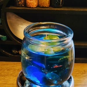 May include: A blue glass jar candle with teal wax and a white wick. The candle is sitting on a silver base and has blue and red glitter inside.