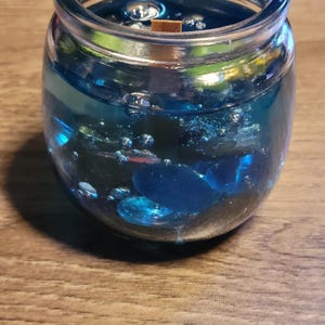 May include: A clear glass jar filled with a blue gel candle with embedded blue and silver glitter. The candle has a wooden wick.