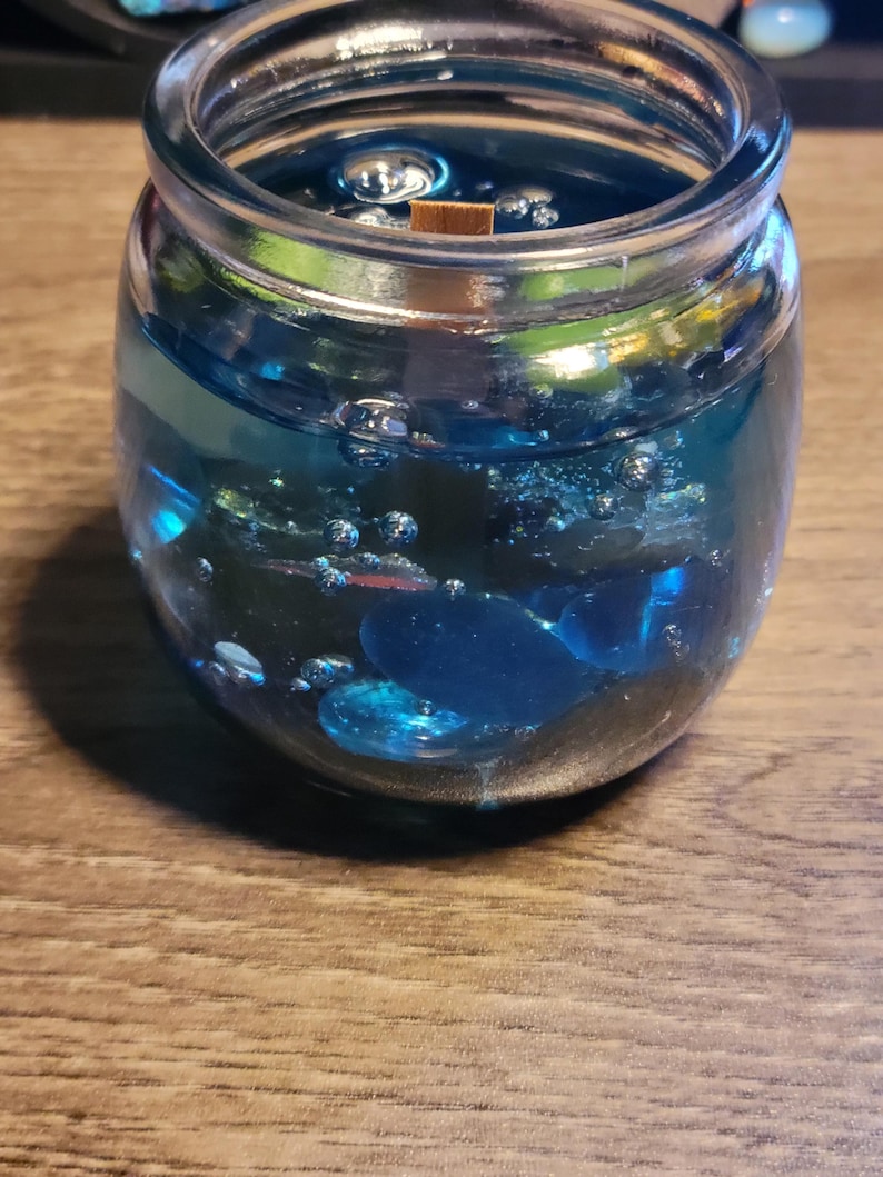 May include: A clear glass jar filled with a blue gel candle with embedded blue and silver glitter. The candle has a wooden wick.