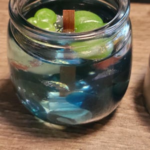 May include: A glass jar candle with a blue wax base and green leaves floating on top. A wooden wick is in the centre of the candle.