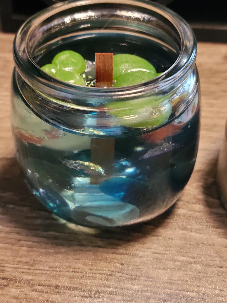 May include: A glass jar candle with a blue wax base and green leaves floating on top. A wooden wick is in the centre of the candle.