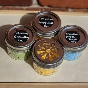 May include: Four small glass jars with metal lids. Three lids have black labels with white text reading Intention: Grounding 4oz, Intention: Happiness 4oz, and Intention: Stress Relief 4oz. One lid has a decorative flower-shaped cutout.