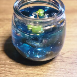May include: A clear glass jar filled with blue liquid and small blue and green glass beads. A small green plant is growing in the liquid.