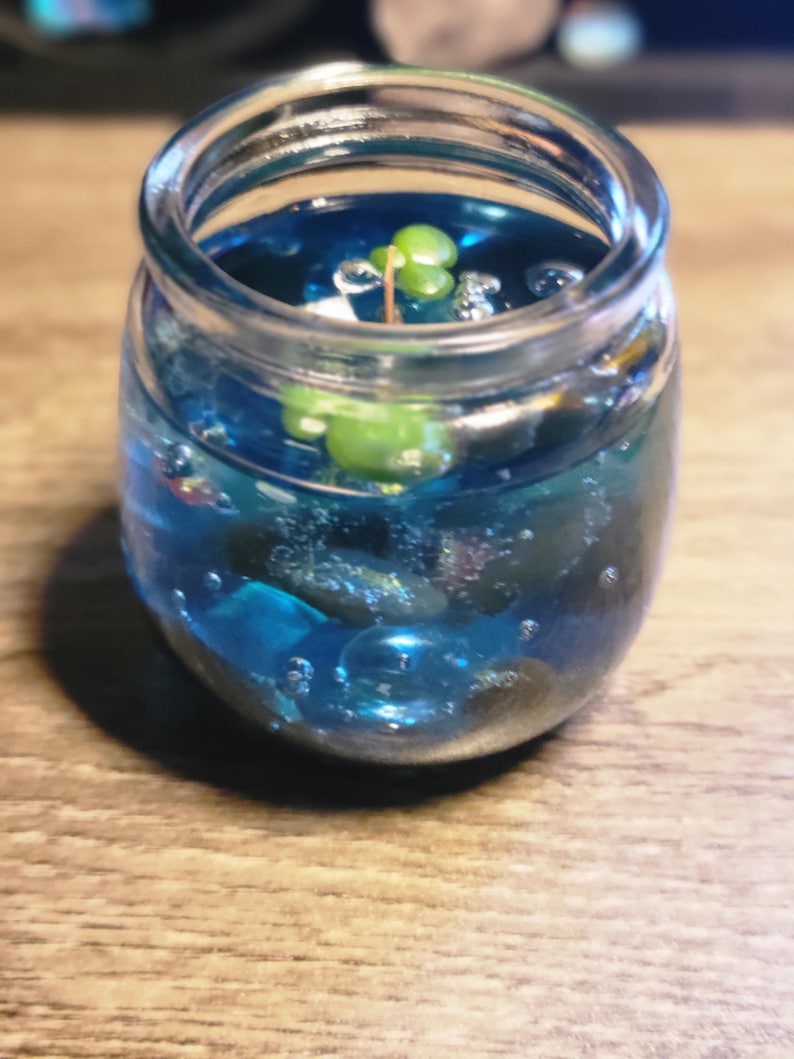 May include: A clear glass jar filled with blue liquid and small blue and green glass beads. A small green plant is growing in the liquid.