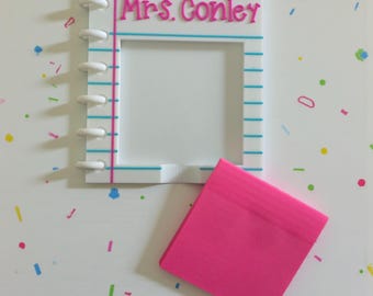 May include: A white acrylic notepad-shaped frame with the name "Mrs. Conley" in pink. The frame has blue lines and a pink spine, with a stack of pink sticky notes next to it. The background is white with colorful confetti.