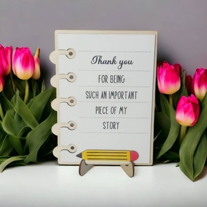 May include: A wooden sign with a white background and black text that reads "Thank you for being such an important piece of my story". The sign is decorated with a yellow pencil and is standing on a small wooden stand. The sign is surrounded by pink tulips.