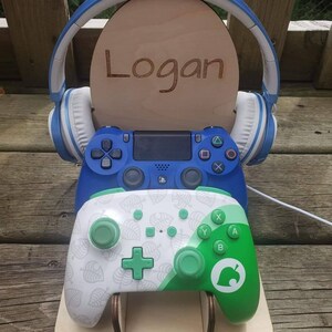 May include: A wooden stand with a blue headset, a blue PlayStation controller, and a green and white Nintendo Switch controller. The stand has a personalized nameplate that says "Logan".
