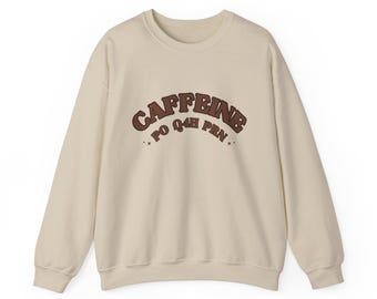 Op de afbeelding: Een beige sweater met ronde hals en de bruine tekst CAFFEINE PO Q4H PRN in een boog over de borst. Het ontwerp bevat kleine steraccenten aan weerszijden van de tekst, die is gedrukt in een vet, afgerond lettertype met een retro uitstraling.