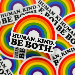 May include: A collection of rainbow-colored stickers with the text "HUMAN. KIND. BE BOTH." The stickers feature a rainbow arch design with the colors red, orange, yellow, green, blue, and purple. The stickers are on a wooden surface.