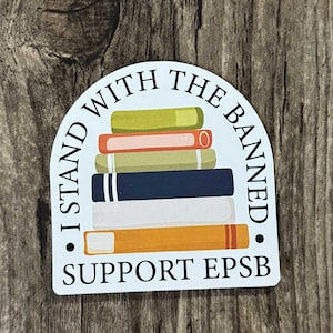 May include: A white sticker featuring a stack of books in various colours, including green, orange, blue, and white. The text reads "I STAND WITH THE BANNED. SUPPORT EPSB" in a semi-circular arrangement.