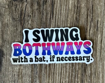 May include: A sticker with the text "I SWING BOTHWAYS with a bat, if necessary." The text is in black, blue, pink, and purple. The sticker has a white border and is on a wooden surface.