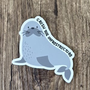 May include: A die-cut sticker featuring a light grey cartoon seal with a darker grey face and flippers. The seal has black whiskers and eyes, and the text CRUSH THE INFRASTRUCTURE is printed in black capital letters along the right side of the seal.