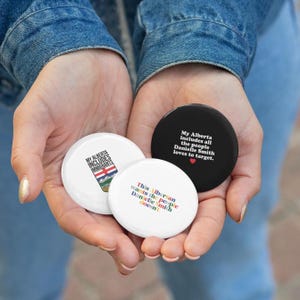 May include: Three round badges are held in open hands. One is black with white text: "My Alberta includes all the people Danielle Smith loves to target." Another is white with the text "My Alberta includes immigrants."