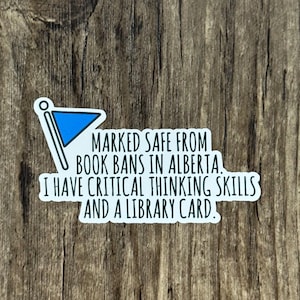 May include: A white sticker with the text "MARKED SAFE FROM BOOK BANS IN ALBERTA. I HAVE CRITICAL THINKING SKILLS AND A LIBRARY CARD." A blue flag graphic is in the upper left corner. The sticker is on a wooden surface.